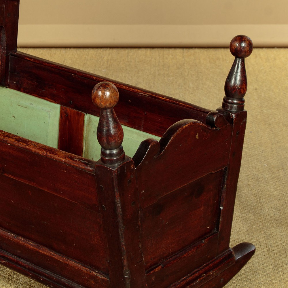 An antique dark brown wooden hooded rocking cradle featuring turned corner finials, a curved rocker base, and a pale green painted interior. View VII