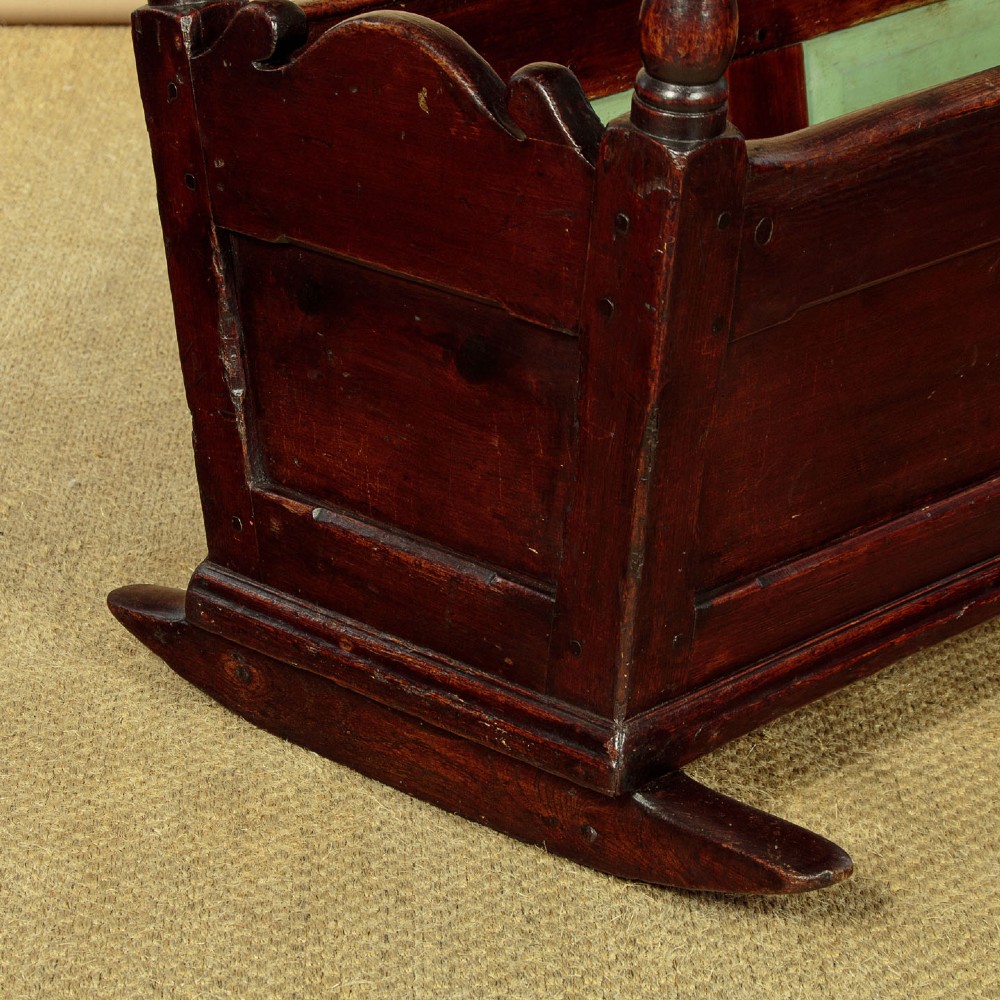 An antique dark brown wooden hooded rocking cradle featuring turned corner finials, a curved rocker base, and a pale green painted interior. View VIII