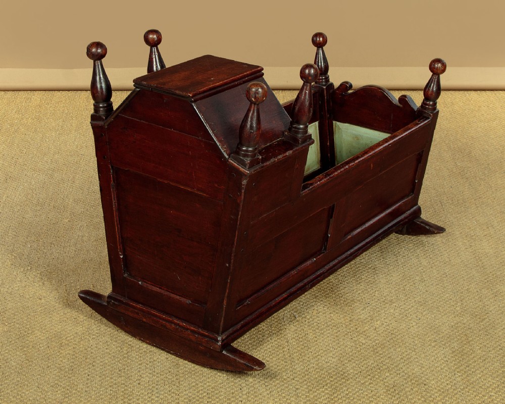An antique dark brown wooden hooded rocking cradle featuring turned corner finials, a curved rocker base, and a pale green painted interior. View XI