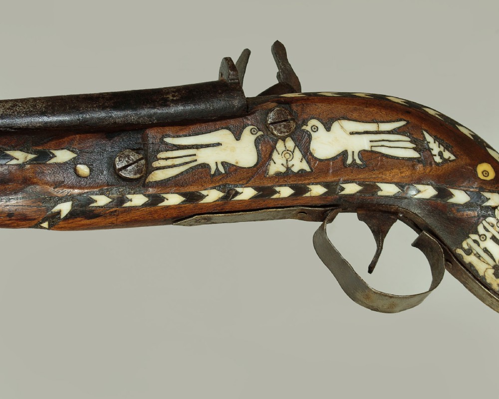 An antique Afghan Jezail musket featuring a distinctly curved wooden stock decorated with geometric bone inlay and a steel lock mechanism. View V