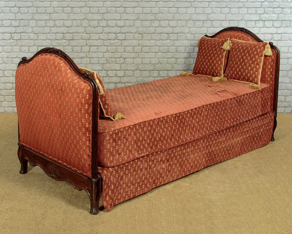 A c.1920 French-style upholstered day bed featuring a dark carved wooden frame, terracotta red patterned fabric, and matching tassel-trimmed cushions. View II