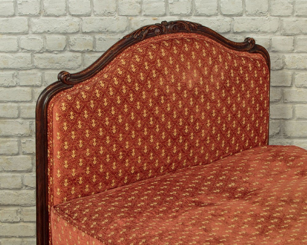 A c.1920 French-style upholstered day bed featuring a dark carved wooden frame, terracotta red patterned fabric, and matching tassel-trimmed cushions. View V