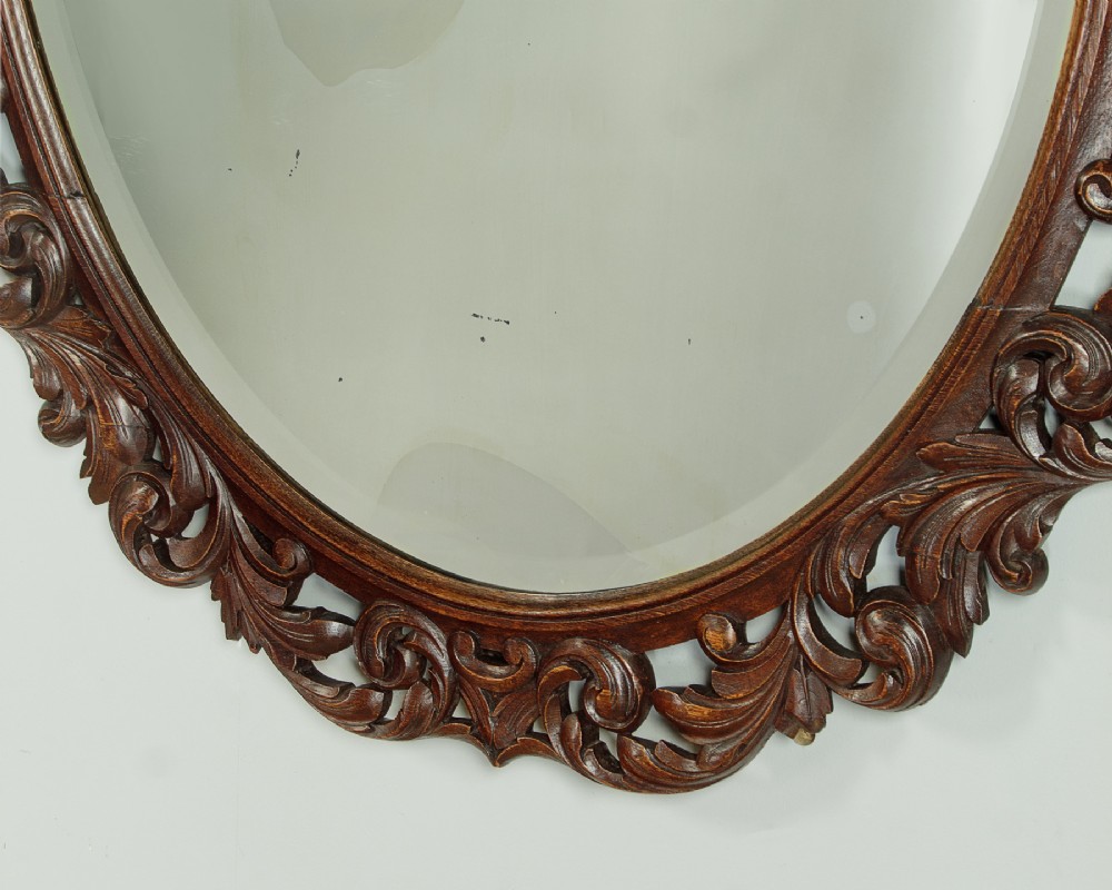 A late Victorian oval wall mirror featuring a carved dark oak frame with acanthus scrollwork and a crest motif enclosing a bevelled glass mirror plate. View VII