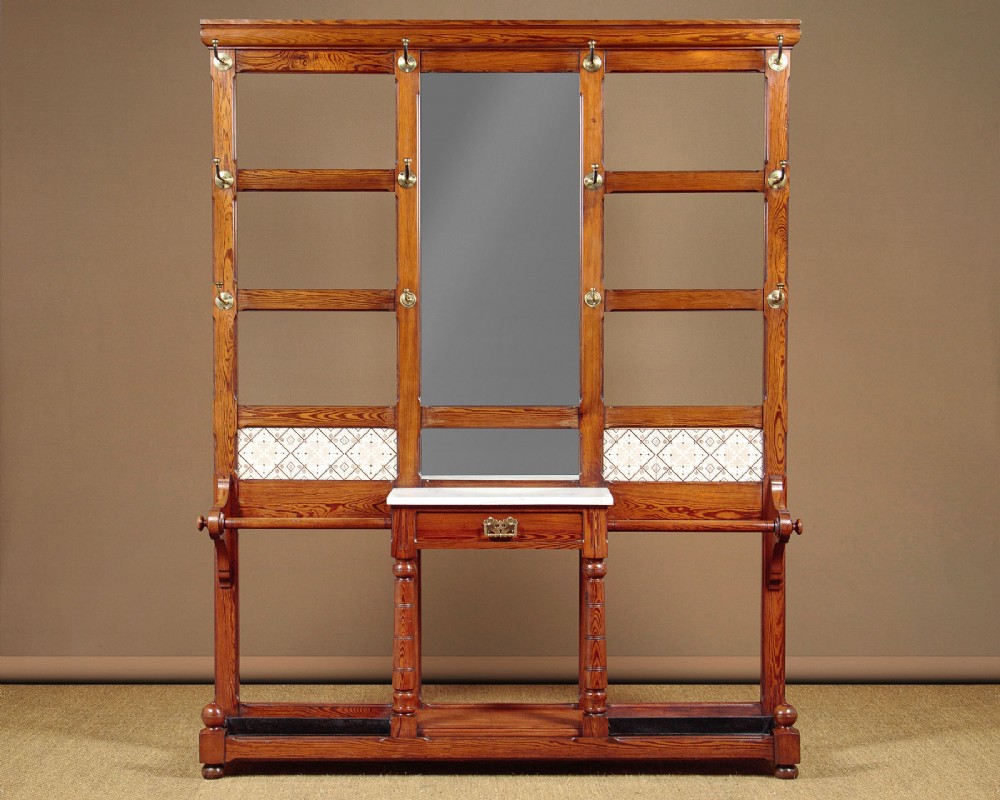 A large Victorian pine hallstand featuring a central vertical mirror, multiple brass coat hooks, inset ceramic tiles, a marble-topped glove drawer, and lower umbrella stand trays.