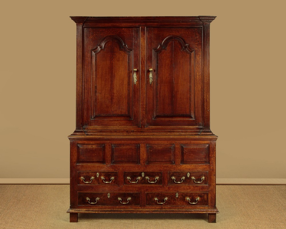 A tall 18th-century dark oak Georgian tack cupboard featuring twin arched panel doors over a base with five drawers and brass handles.