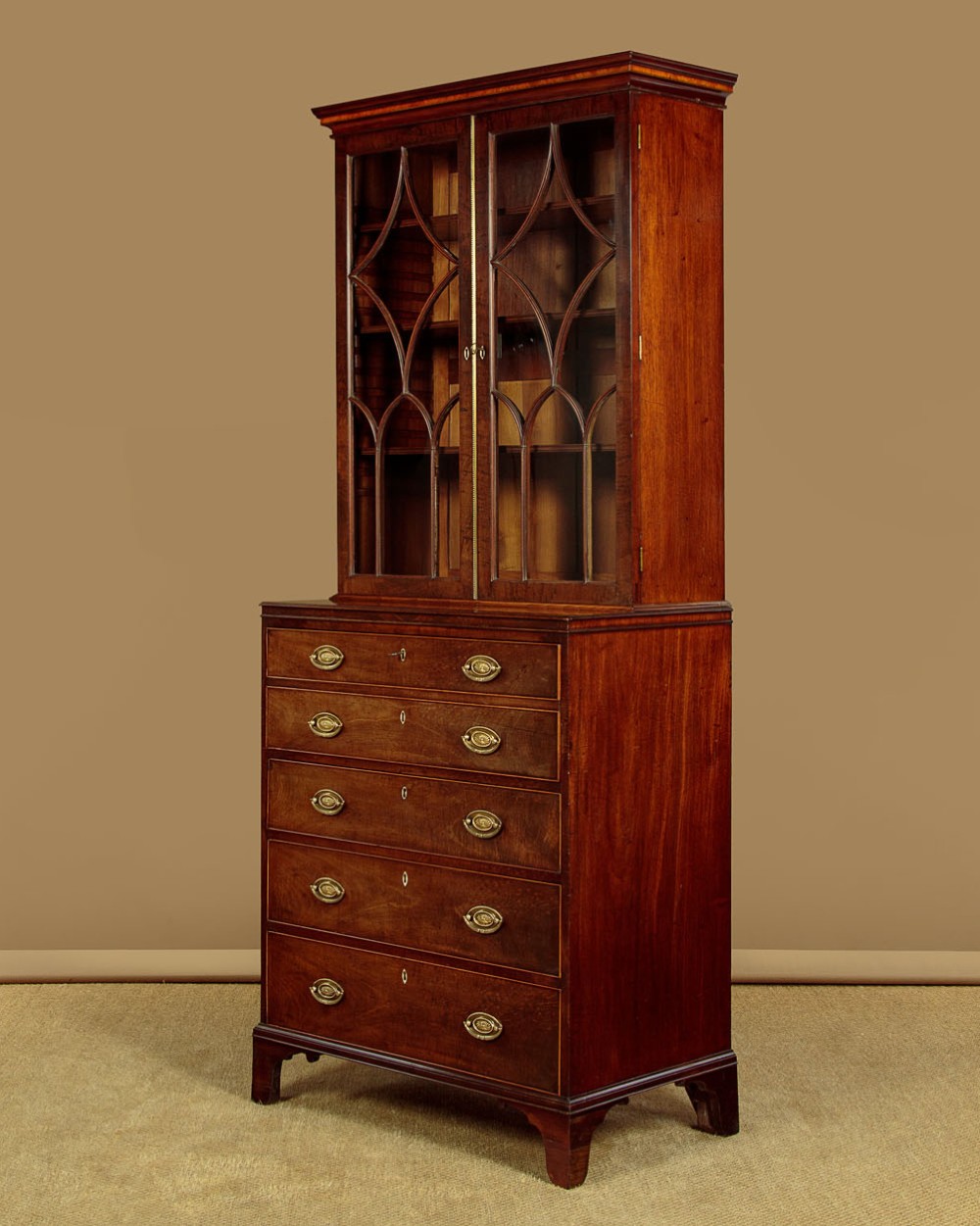 A tall mahogany secretaire bureau bookcase featuring two upper glazed doors with geometric astragal glazing over a chest of drawers with brass oval handles. View IV