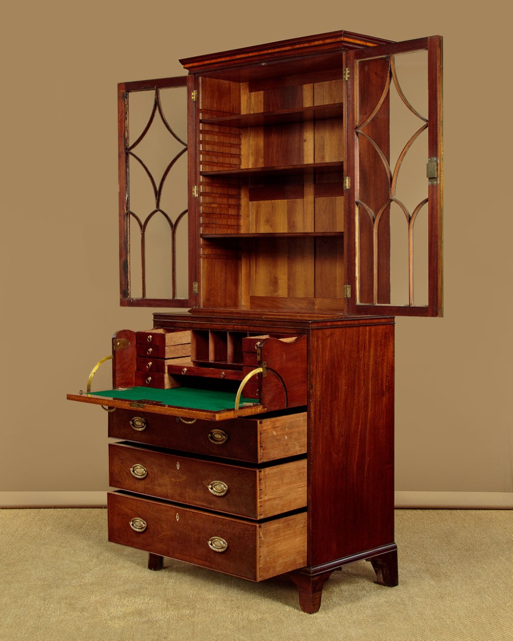 A tall mahogany secretaire bureau bookcase featuring two upper glazed doors with geometric astragal glazing over a chest of drawers with brass oval handles. View V