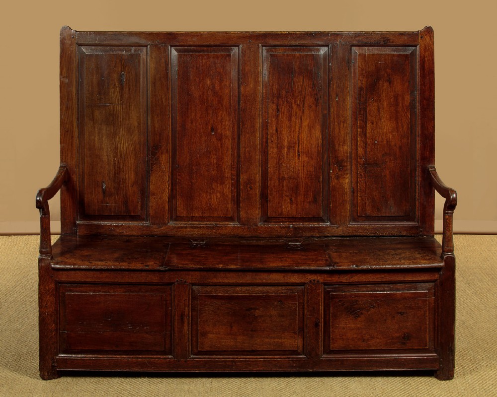 A dark oak 18th-century high-back box settle featuring fielded panels, shaped open armrests, and a hinged lid seat.