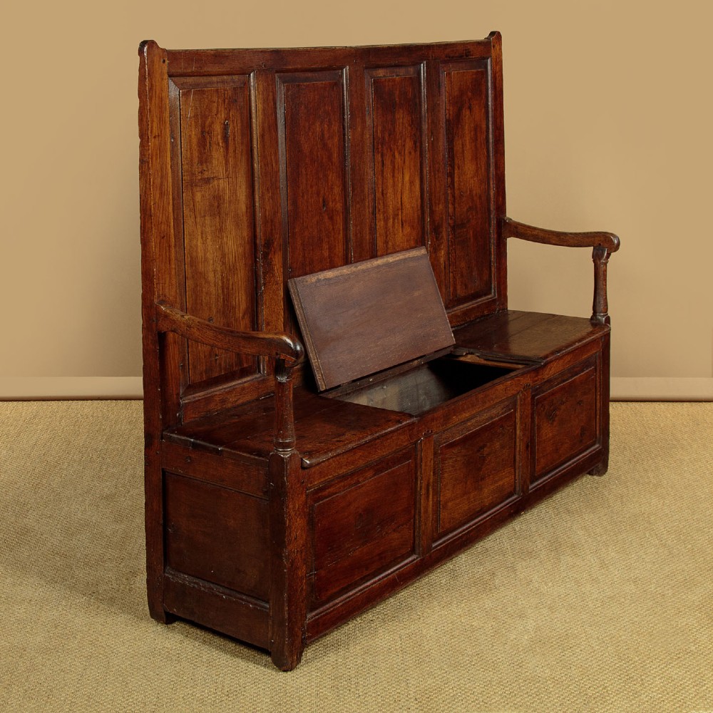 A dark oak 18th-century high-back box settle featuring fielded panels, shaped open armrests, and a hinged lid seat. View IV