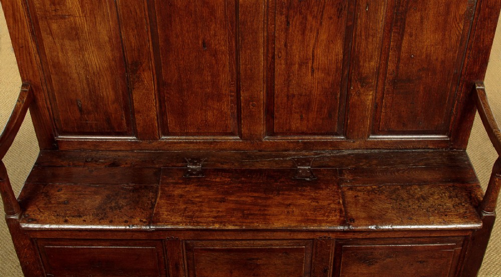A dark oak 18th-century high-back box settle featuring fielded panels, shaped open armrests, and a hinged lid seat. View VI