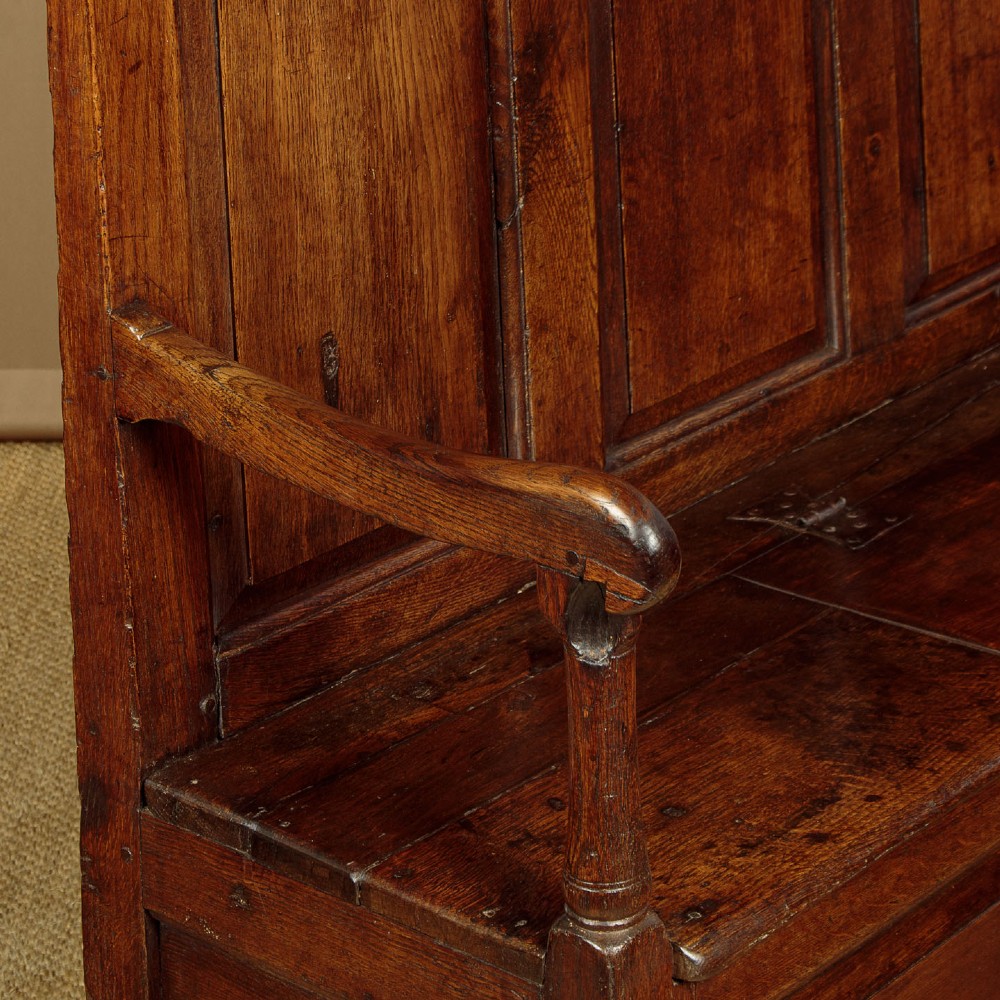 A dark oak 18th-century high-back box settle featuring fielded panels, shaped open armrests, and a hinged lid seat. View VII