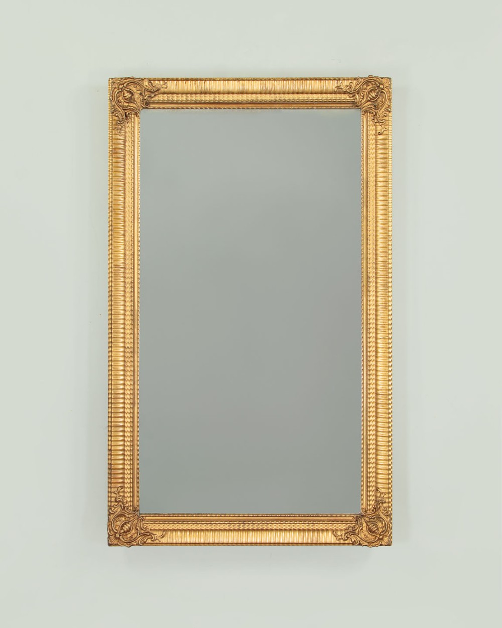 A rectangular wall mirror featuring a gilded frame with reeded moulding and carved scrollwork details in each corner.