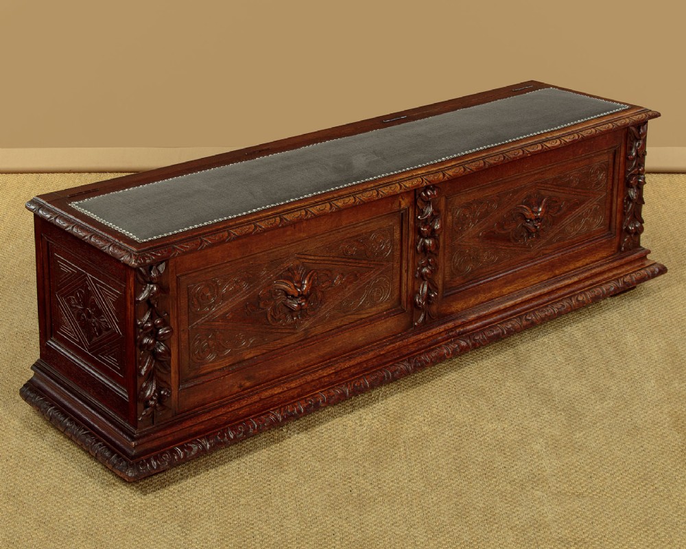 A long late Victorian dark oak ottoman box seat featuring an inset leather top with brass studding, carved relief mask panels, and a heavy moulded base.