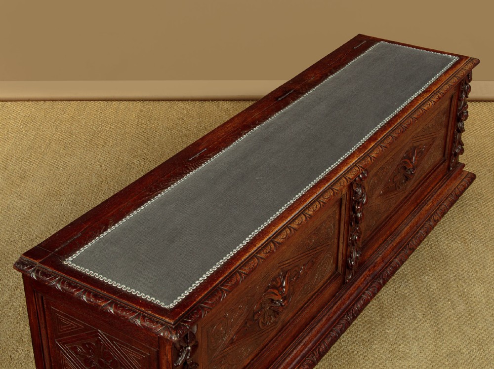 A long late Victorian dark oak ottoman box seat featuring an inset leather top with brass studding, carved relief mask panels, and a heavy moulded base. View IV