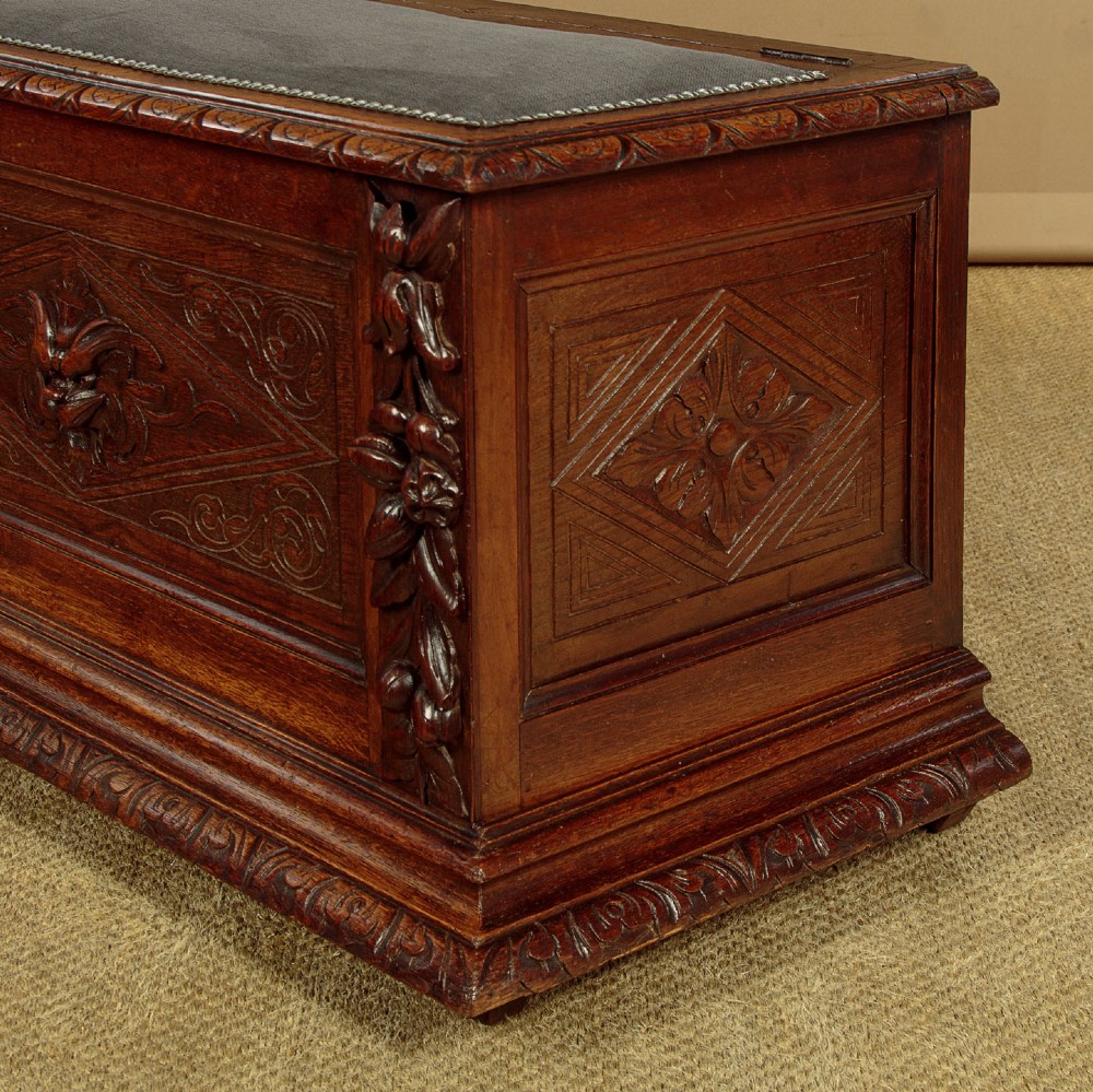 A long late Victorian dark oak ottoman box seat featuring an inset leather top with brass studding, carved relief mask panels, and a heavy moulded base. View IX