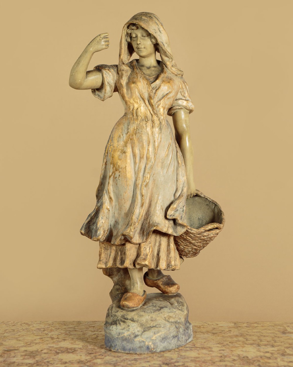 A turn-of-the-century terracotta sculpture depicting a standing young woman in rural dress wearing a headscarf and clogs, holding a basket.