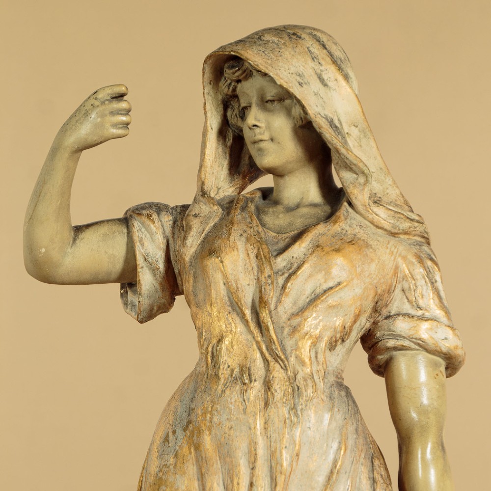 A turn-of-the-century terracotta sculpture depicting a standing young woman in rural dress wearing a headscarf and clogs, holding a basket. View IV
