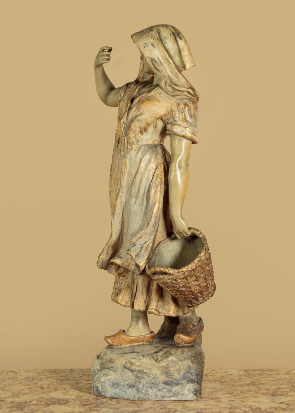 A turn-of-the-century terracotta sculpture depicting a standing young woman in rural dress wearing a headscarf and clogs, holding a basket. View V