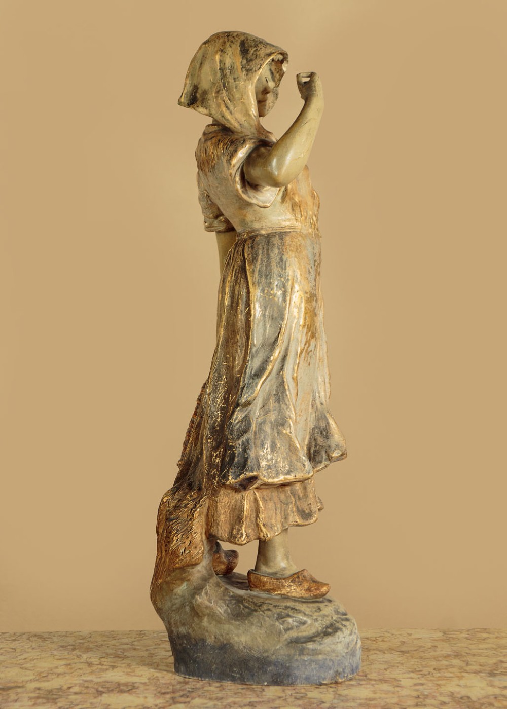 A turn-of-the-century terracotta sculpture depicting a standing young woman in rural dress wearing a headscarf and clogs, holding a basket. View VI
