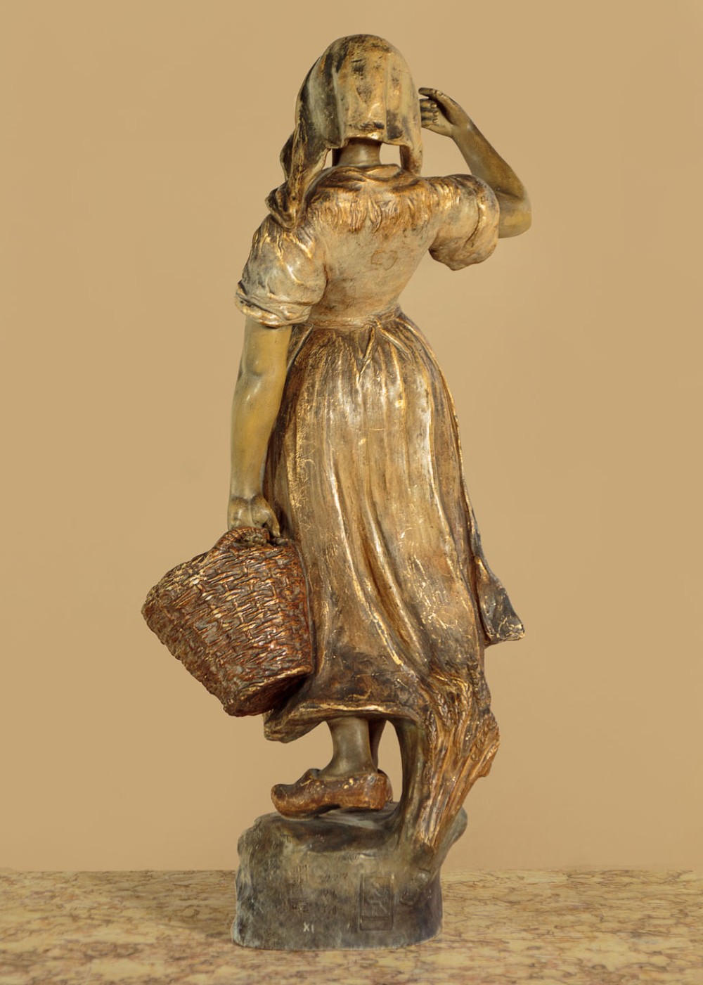A turn-of-the-century terracotta sculpture depicting a standing young woman in rural dress wearing a headscarf and clogs, holding a basket. View VII