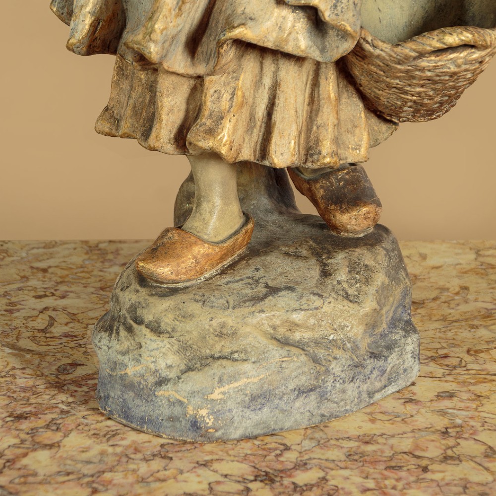 A turn-of-the-century terracotta sculpture depicting a standing young woman in rural dress wearing a headscarf and clogs, holding a basket. View VIII