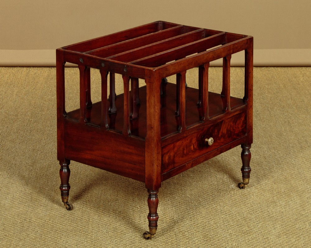 A dark mahogany Canterbury featuring four open slatted dividers above a single drawer with turned legs and brass castors.