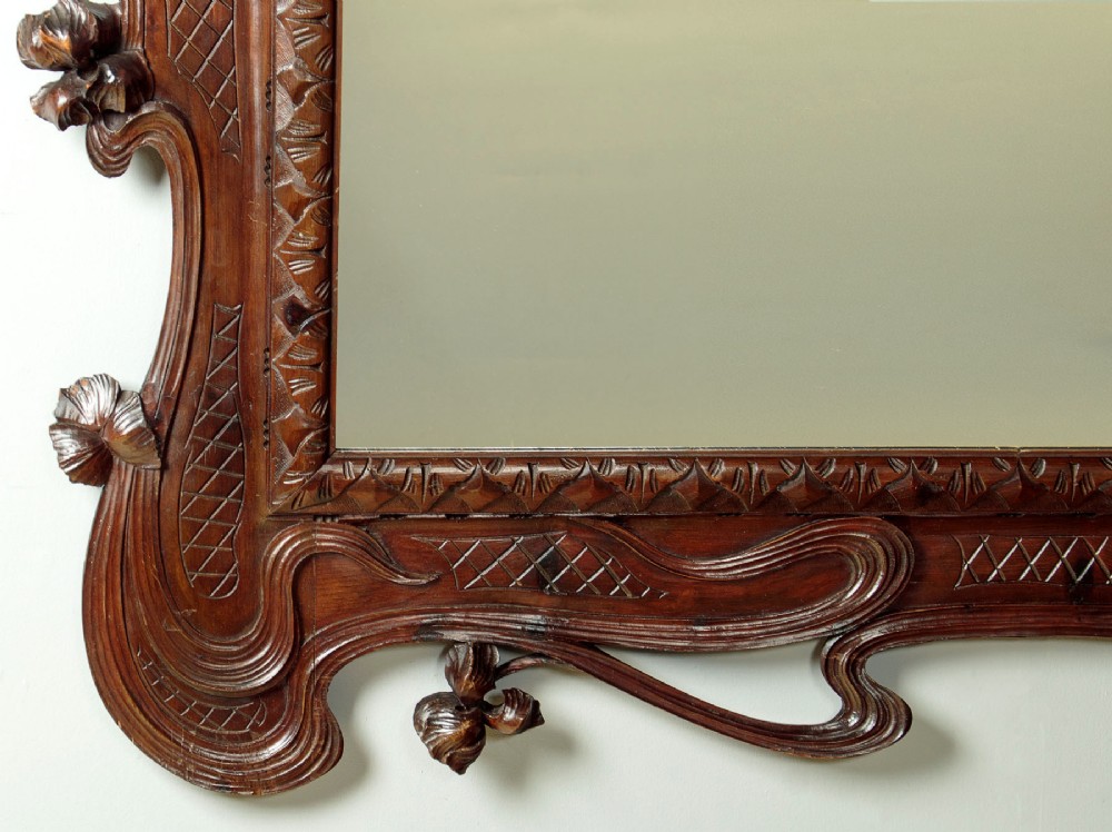 An antique Art Nouveau wall mirror featuring an intricately carved dark wood frame with sweeping whiplash curves and floral accents surrounding rectangular mirror glass. View VI