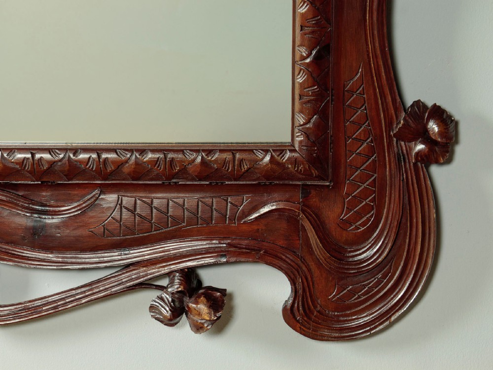 An antique Art Nouveau wall mirror featuring an intricately carved dark wood frame with sweeping whiplash curves and floral accents surrounding rectangular mirror glass. View VII