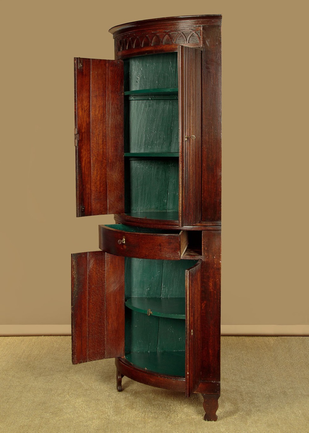 A tall early 19th-century dark mahogany double height corner cupboard featuring reeded front doors, a central drawer, and Gothic arcading at the cornice. View III