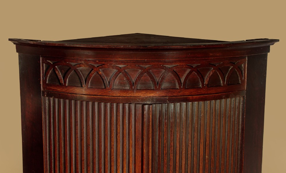 A tall early 19th-century dark mahogany double height corner cupboard featuring reeded front doors, a central drawer, and Gothic arcading at the cornice. View IV