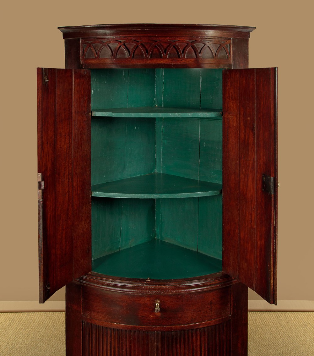 A tall early 19th-century dark mahogany double height corner cupboard featuring reeded front doors, a central drawer, and Gothic arcading at the cornice. View VI