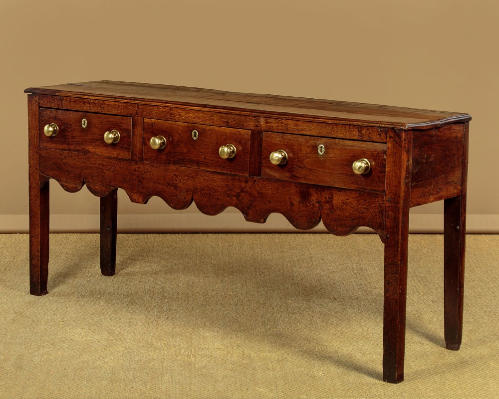An 18th-century oak dresser base featuring three drawers with brass knobs, a scalloped lower apron, and four straight legs.