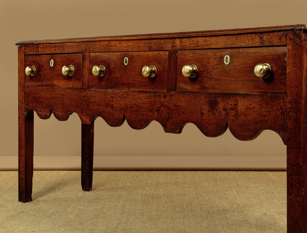 An 18th-century oak dresser base featuring three drawers with brass knobs, a scalloped lower apron, and four straight legs. View VII