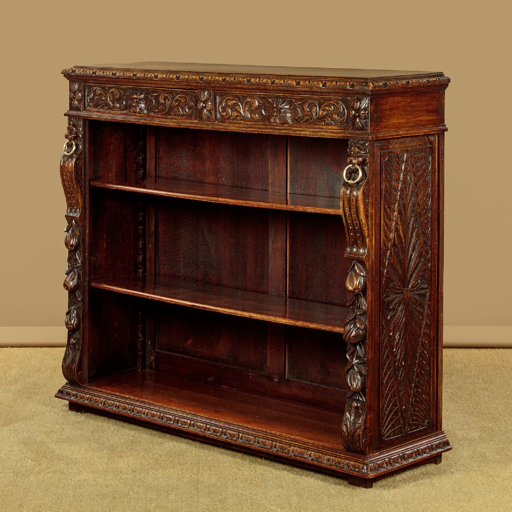 A low late Victorian dark oak open bookcase with detailed foliate relief carvings, brass ring fittings on side pilasters, and adjustable shelves.