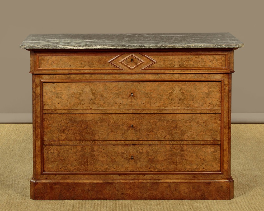 A nineteenth-century burr walnut commode featuring a grey marble top surface, a upper drawer with a central diamond inlay, three long lower drawers, and a plinth base.