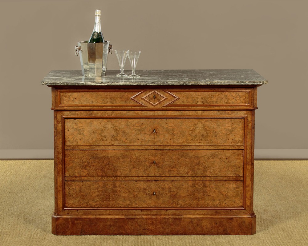 A nineteenth-century burr walnut commode featuring a grey marble top surface, a upper drawer with a central diamond inlay, three long lower drawers, and a plinth base. View II