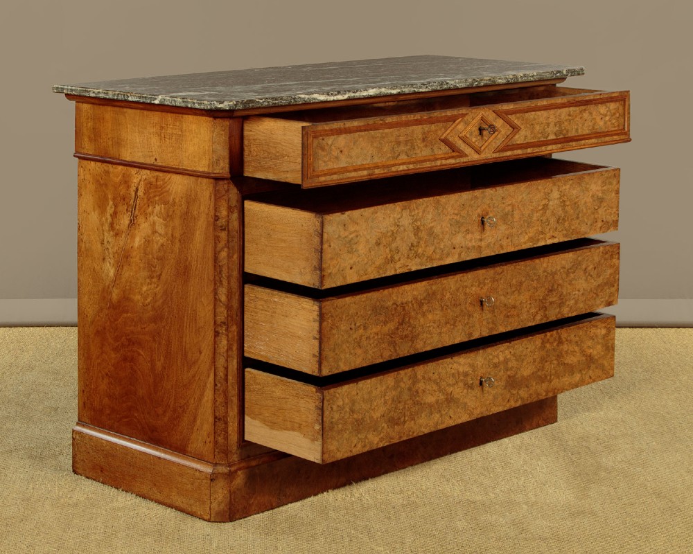 A nineteenth-century burr walnut commode featuring a grey marble top surface, a upper drawer with a central diamond inlay, three long lower drawers, and a plinth base. View V