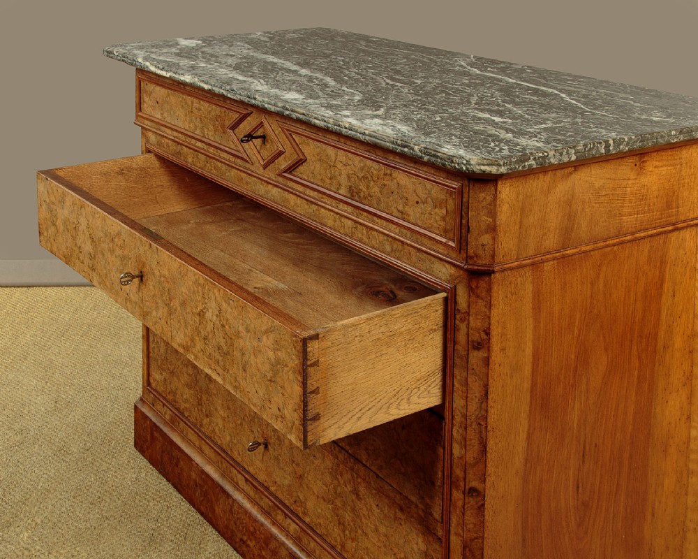 A nineteenth-century burr walnut commode featuring a grey marble top surface, a upper drawer with a central diamond inlay, three long lower drawers, and a plinth base. View XI