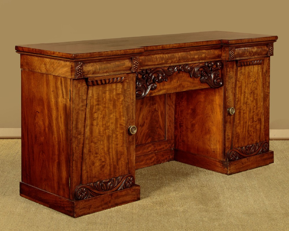 An early Victorian flame mahogany pedestal sideboard featuring carved scrollwork across the central apron and pedestal cupboards set on plinth bases.