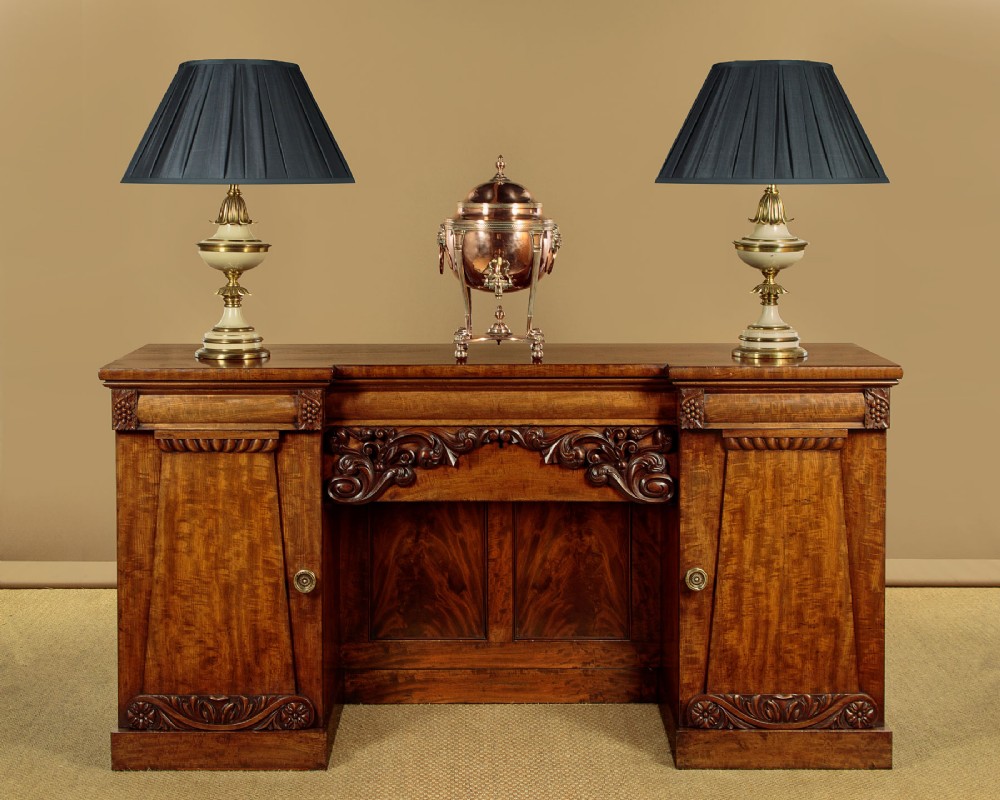 An early Victorian flame mahogany pedestal sideboard featuring carved scrollwork across the central apron and pedestal cupboards set on plinth bases. View II