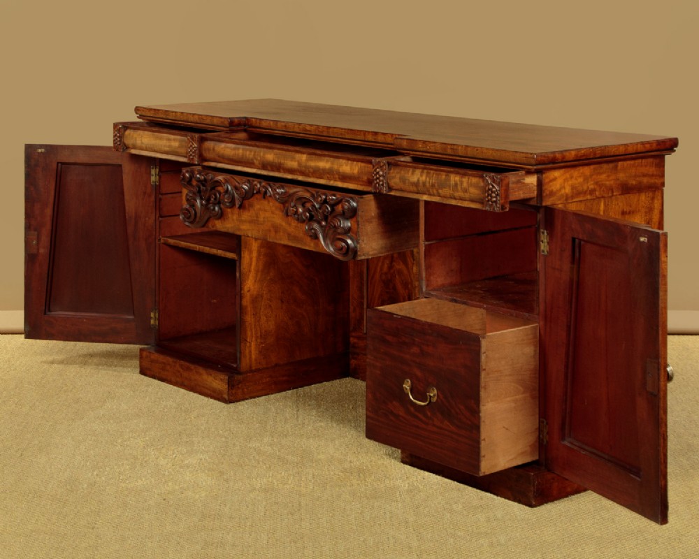 An early Victorian flame mahogany pedestal sideboard featuring carved scrollwork across the central apron and pedestal cupboards set on plinth bases. View IV