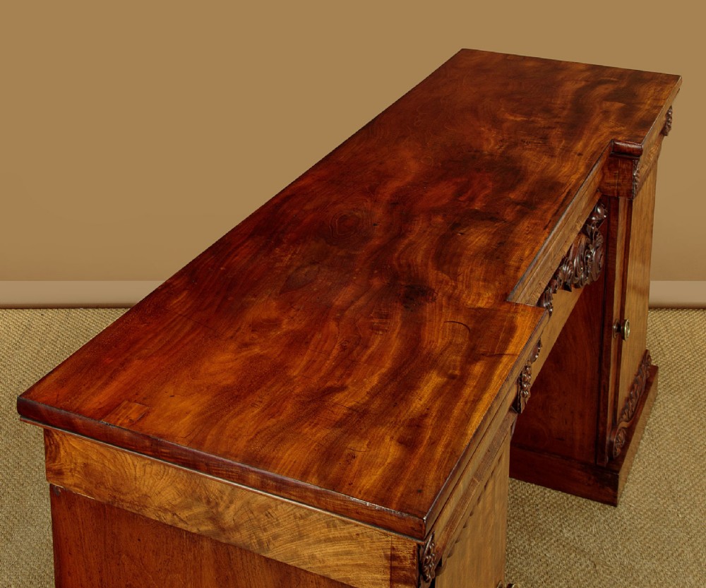 An early Victorian flame mahogany pedestal sideboard featuring carved scrollwork across the central apron and pedestal cupboards set on plinth bases. View V
