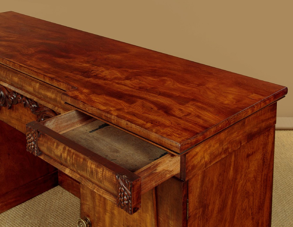 An early Victorian flame mahogany pedestal sideboard featuring carved scrollwork across the central apron and pedestal cupboards set on plinth bases. View VI