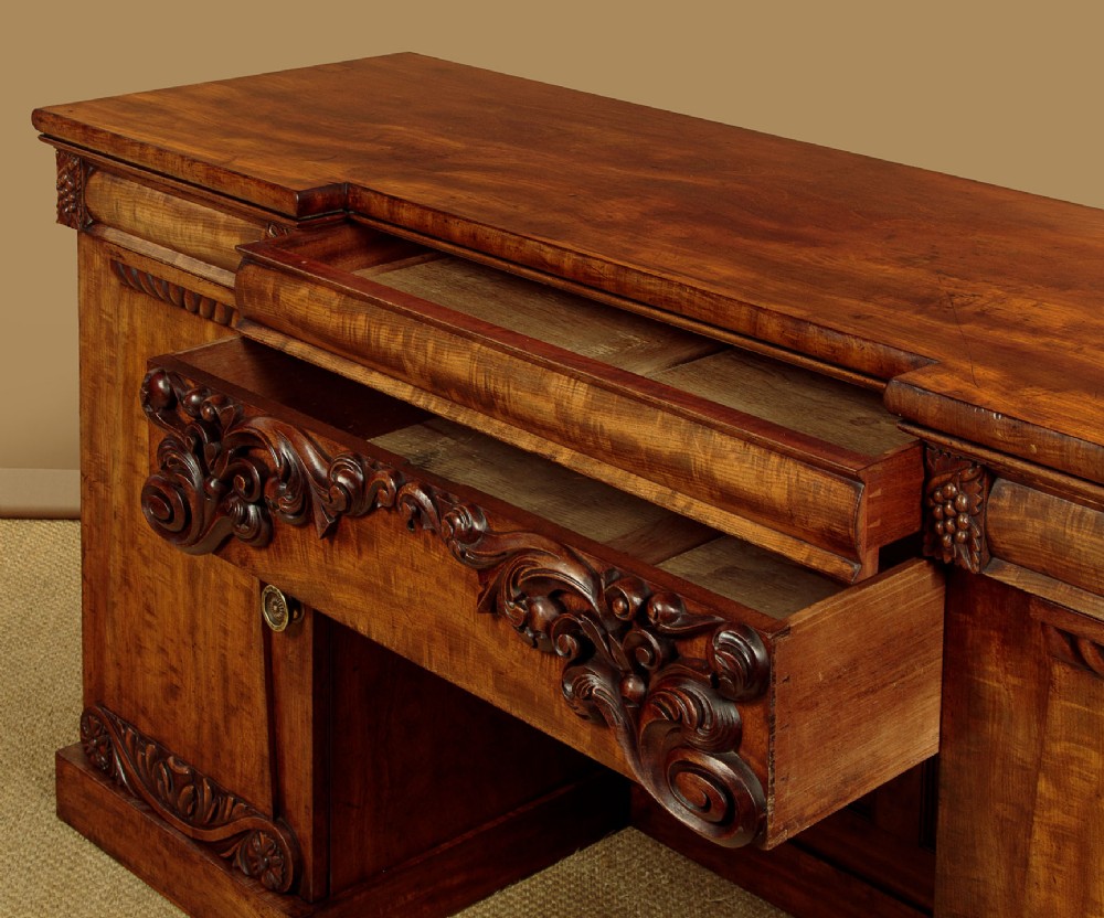 An early Victorian flame mahogany pedestal sideboard featuring carved scrollwork across the central apron and pedestal cupboards set on plinth bases. View VII