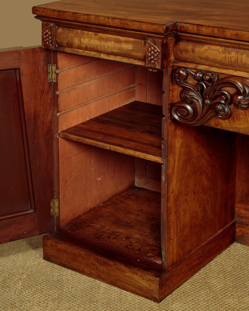 An early Victorian flame mahogany pedestal sideboard featuring carved scrollwork across the central apron and pedestal cupboards set on plinth bases. View X