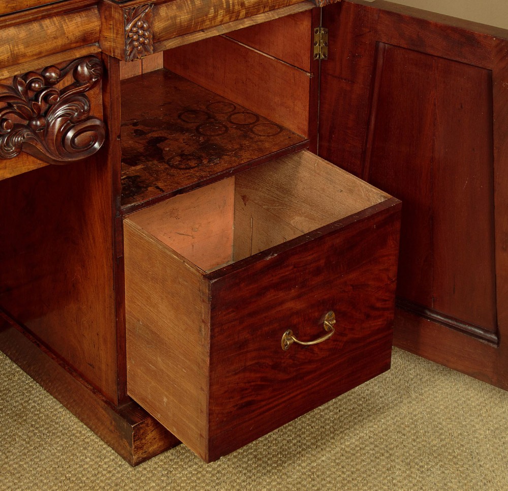 An early Victorian flame mahogany pedestal sideboard featuring carved scrollwork across the central apron and pedestal cupboards set on plinth bases. View XI