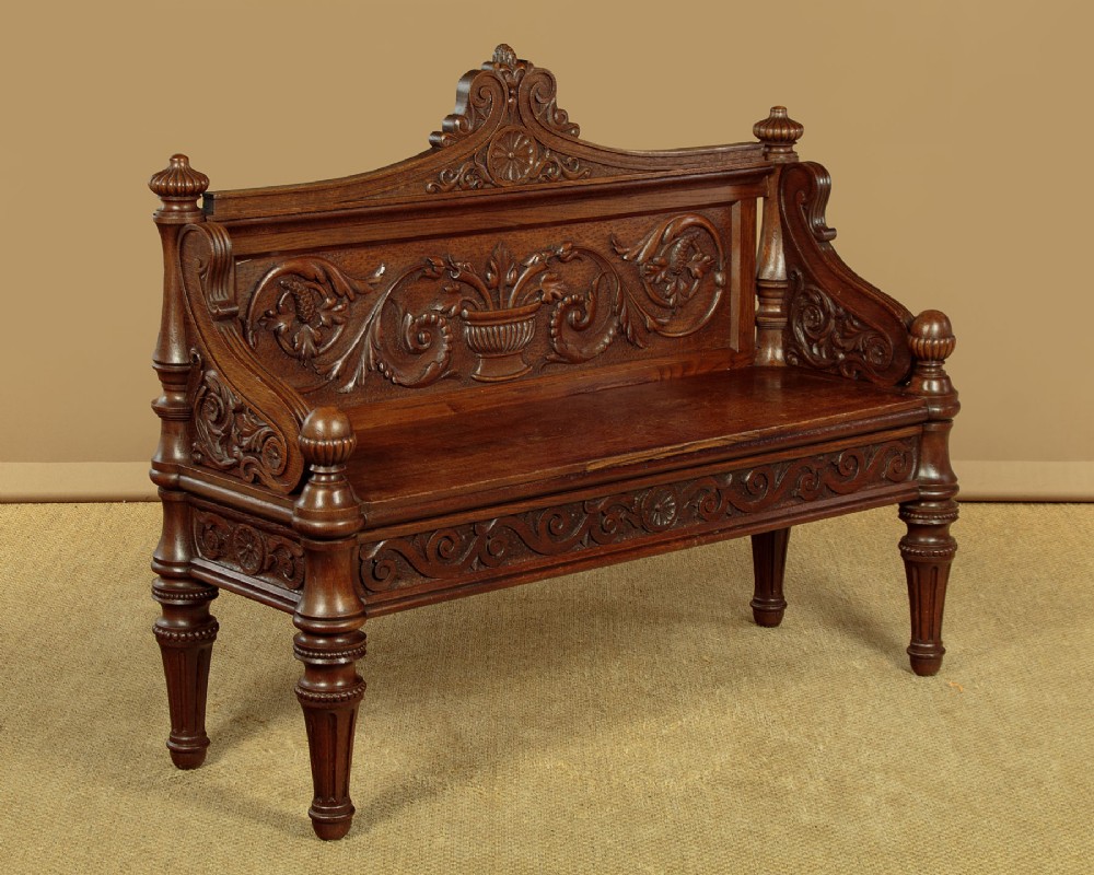 A carved dark oak hall bench featuring a scroll decorated backrest, carved relief panels with an urn motif, turned fluted front legs, and shaped side armrests.