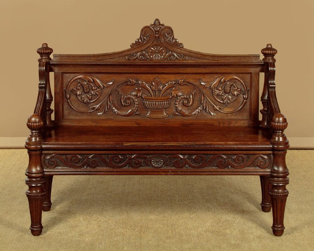 A carved dark oak hall bench featuring a scroll decorated backrest, carved relief panels with an urn motif, turned fluted front legs, and shaped side armrests. View II