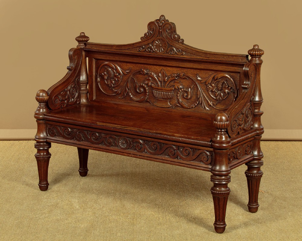 A carved dark oak hall bench featuring a scroll decorated backrest, carved relief panels with an urn motif, turned fluted front legs, and shaped side armrests. View III