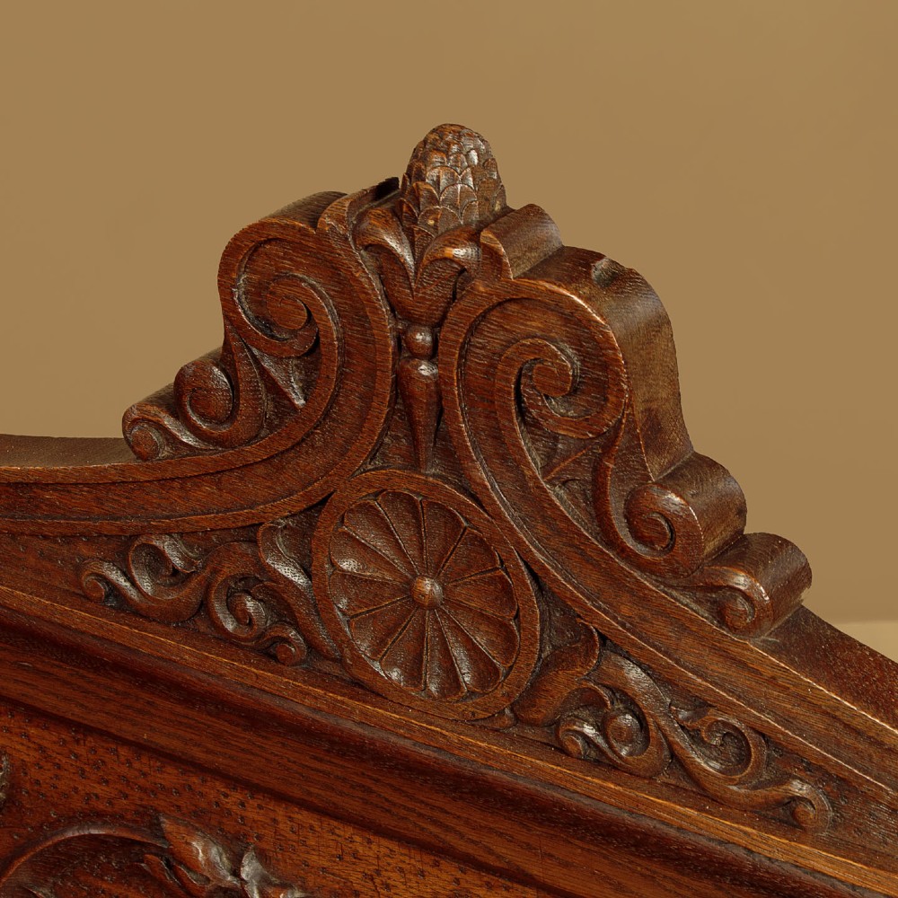 A carved dark oak hall bench featuring a scroll decorated backrest, carved relief panels with an urn motif, turned fluted front legs, and shaped side armrests. View IV